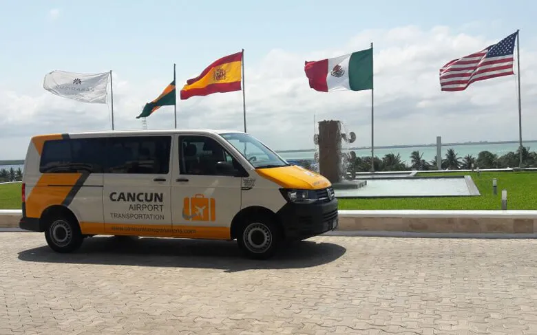 Van labeled with Cancun Airport Transportation corporate image parked in front of a water fountain located at Paradisus Resort with several flags, including United States, Spain and Mexico flags. A lagoon, blue sky and some clouds on the background.